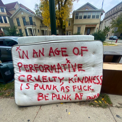 Red spray paint on a white mattress: "In an age of performative cruelty, kindness is punk as fuck. Be punk AF."
