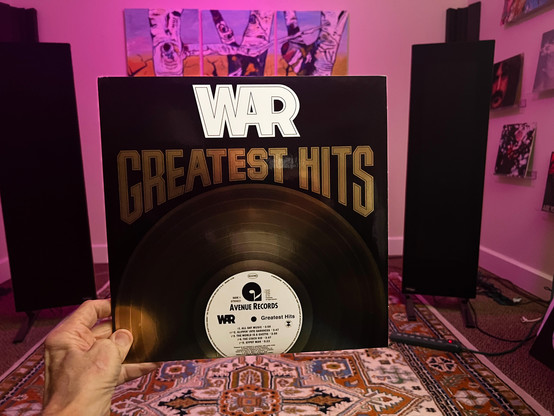 Me holding the album WAR, Greatest Hits. In the background are my speakers