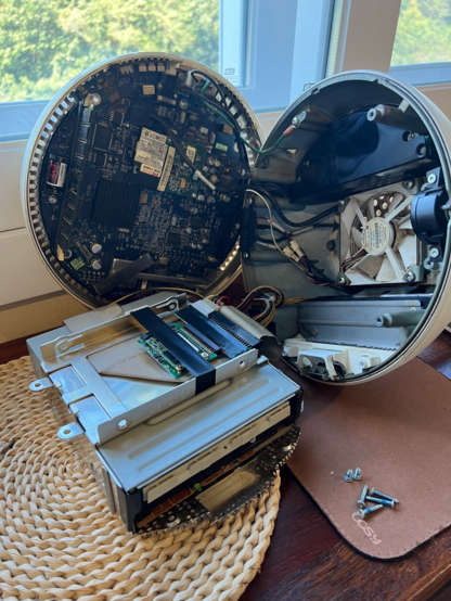 Photo of the iMac G4 opened up, with the main body laying to the right, the bottom part laying up against the window frame, and the disc and "hard drive" caddy laying in front. There is no actual hard drive installed anymore, having been replaced by me with an IDE to mSATA adapter. 