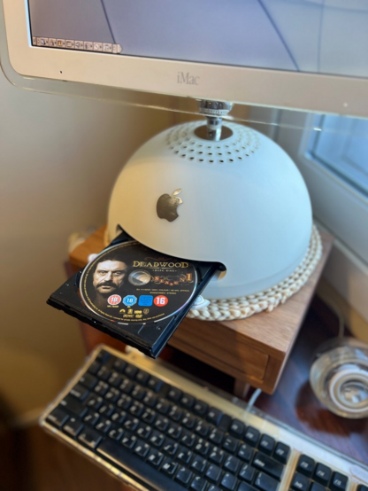 Photo showing the iMac G4 all put back together and its disc tray opened with a DVD of the HBO show Deadwood inside. The disc tray is black, which is different from the stock drive's white color, but it doesn't look out of place at all, with the other inside parts visible when the tray is open also being black, as-well as the keyboard keys being black too.
