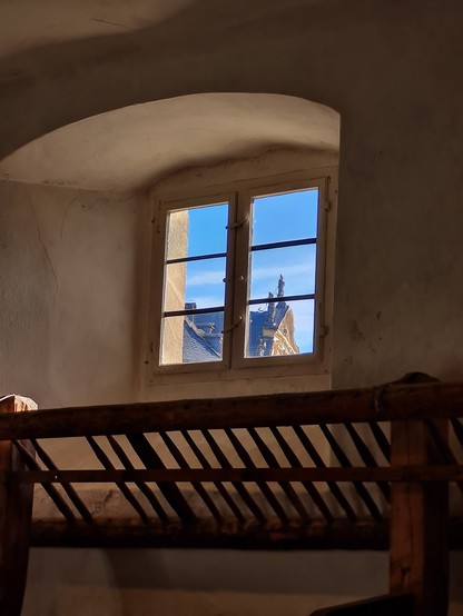 Blick auf ein historisches Fenster. Draußen zu sehen der Giebel der neuen Residenz Bamberg. Innen eine Futtertraufe eines ehemaligen Pferdestalls 