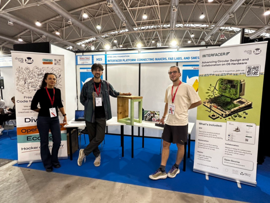 Three people standing in front of a exhibition booth with rollups and a self-built chair.