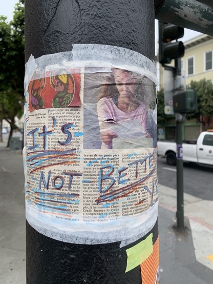 A piece of newspaper taped to a post on Valencia street with masking tape. The artist has written "It's not better yet" in blue ink and paint pens. There is also a cut up from a book that shows a drawing of some lungs with pieces of food in them. 