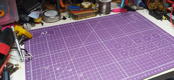 A messy desk where everything has been pushed back to allow a new purple A3 cutting mat to be placed. 