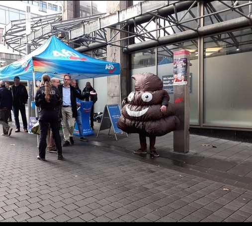 Typ in Scheißehaufen Verkleidung steht neben einem AfD Stand 