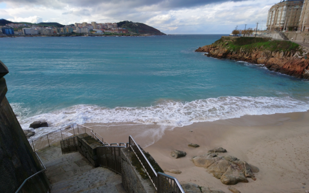Vista desde el paseo marítimo de la playa del Matadero, en Coruña. El océano tiene un color azul celeste, la playa está vacía y a la izquierda, en la parte inferior, se ven las escaleras que nos llevan a dicha playa.

Al fondo se ve el monte de San Pedro, Labañou y algunos de los edificios del barrio de los Rosales bajo un cielo gris azulado a causa de las nubes.
