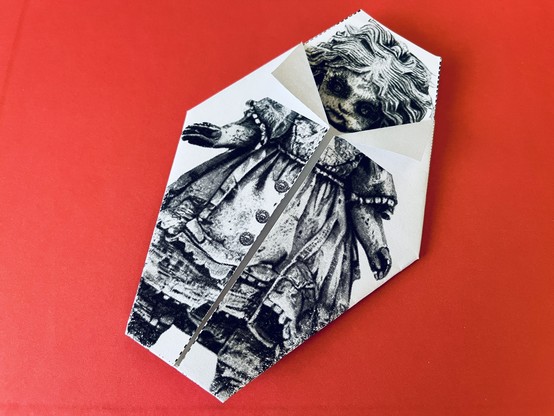 An old and creepy-looking origami doll with intricate details and big, haunting eyes, set against a red background.