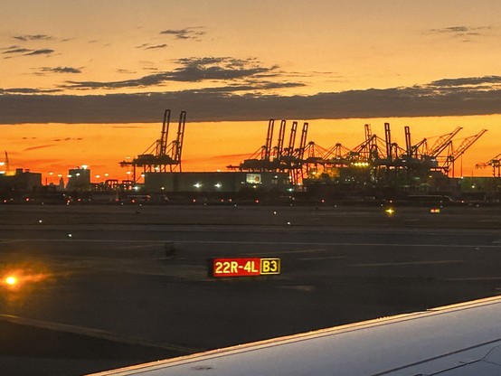 Photo of sunrise with some cranes in the background (the type that unload ships) and an airplane wing in the foreground. 