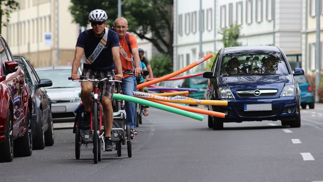 Mehrere Personen fahren im Rahmen einer Fahrraddemo hintereinander mit dem Fahrrad auf einer Straße. An den Fahrrädern sind Poolnudeln befestigt, die vom Fahrrad abstehen und den nötigen Überholabstand markieren. Die Gruppe der Radfahrenden wird von einem Auto mit Mindestabstand überholt.