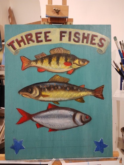 A portrait-orientated board, painted with a blue green background, and three brightly colored fish one above the other. A perch, a brown trout, and a roach. The top has a banner exclaiming "THREE FISHES". At the bottom are two blue stars awaiting embellishment.