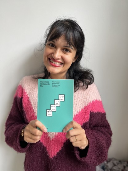 Asian woman smiling holding a copy of a teal coloured book entitled Machine Readable Me, with pixel art icons on the cover, against a white background. 