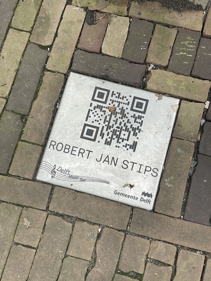 Metalen tegel in een bakstenen straat, met daarop de naam ROBERT JAN STIPS en een QR code die wijst naar de link https://delftmusictour.nl/29-robert-jan-stips/
