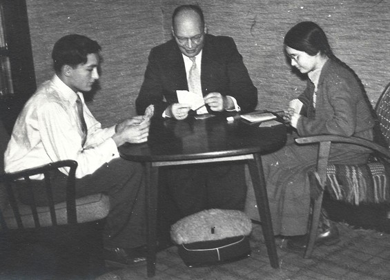 black and white family portrait of a father playing a card game, dated 1957. They are sitting around a square(ish) table, father at the back, son on the left, daughter on the right. Men are wearing white shirts and ties for christmas, father also a dark jacket or suit. Contrasting with this outfit, father's feet are in a fluffy bag to keep them warm. 