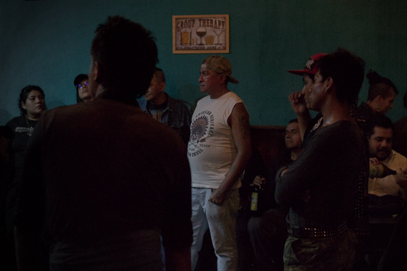 Fotografía de un grupo de personas escuchando un grupo de punk al fondo se observa un cartel en inglés sobre terapia de grupo y unas copas dibujadas 