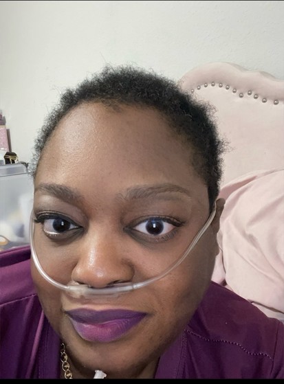 Photo of me, Tinu Abayomi-Paul , A Black Disabled woman with a newly short Afro, smiling slightly at the camera with lips closed, in a purple shirt and purple lipstick and penciled eyebrows but no other makeup.