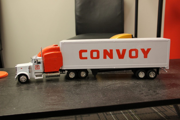 A toy freight truck sits on a dark desk surface. The name "Convoy" is written in big block lettering down the side of the trailer.