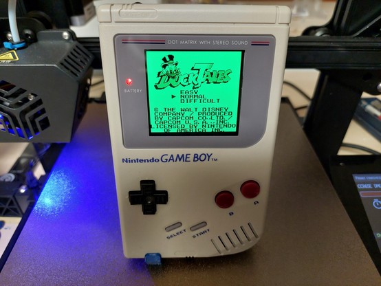 A Nintendo Gameboy DMG01 standing on a PEI print bed of a Creality CR6-SE. The Gameboy is switched on and you can see the title screen of the game Ducktales.