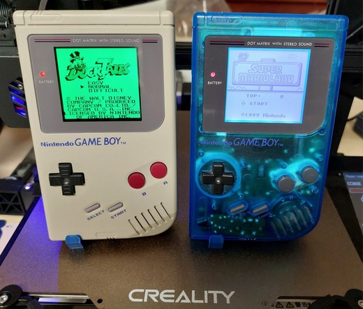 Two Nintendo Gameboy DMG01 (one in the usual grey color and one in transparent blue plastic) standing on the printing bed of a Creality CR6-SE printer. Both Gameboys are running games, the left runs Ducktales and the right one shows the title screen of Super Mario Land.