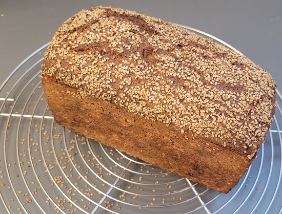 Farbfoto: Das beschriebene Kastenbrot auf dem Kuchengitter. Die Oberfläche ist mit Sesam bestreut und leicht eingerissen. Rundherum liegt auch ziemlich viel Sesam rum. 