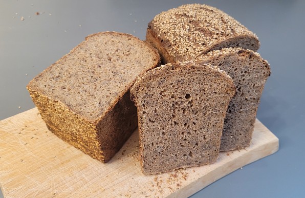 Farbfoto: Dasselbe Brot auf dem Schneidebrett, teilweise aufgeschnitten, so dass man die mittelporige Krume mit ein paar Leinsamenkörnern sehen kann. Die Unterseite und Oberseite des Brotes sind mit Sesam bestreut. 