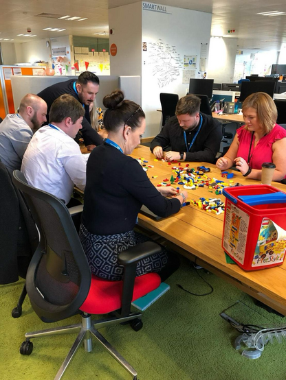 Group of people sat at wooden table, building models out of LEGO 