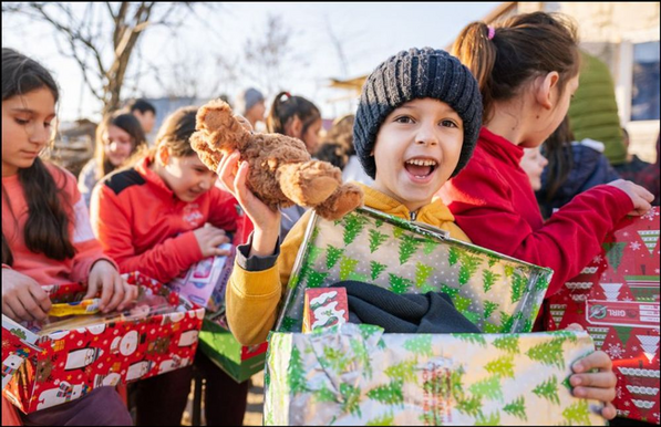 Auf dem Foto sind viele Kinder mit teilweise geöffneten Kartons verpackt mit Weihnachtspapier zu sehen. Im Vordergrund steht ein Kind mit einem geöffneten Karton und in der einen Hand hält es einen Teddy und lacht mit geöffnetem Mund in die Kamera. Das Foto stammt von „Weihnachten im Schuhkarton“