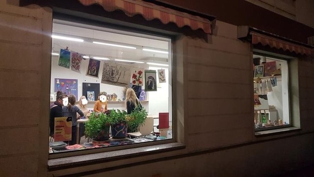 Außenansicht vom LADEN in Wurzen mit zwei hell erleuchteten Fenstern. Innen sieht man Kinder und Erwachsene an einem Tisch und ringsrum viele bunte Bilder.