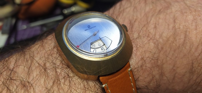 A gold coloured cased watch on a brown leather strap on my wrist. The face is brushed silver metal and has one second hand with two dials for the hours and minutes in the "jump hour" style. 