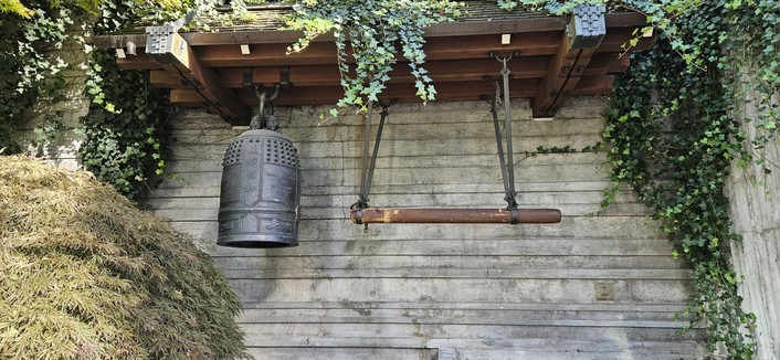 The Japanese friendship bell. The bell is cylindrical and dull grey, on the left. A wooden hammer that strikes it is on the right. Both are underneath a wooden awning. Green plants surround the area.
