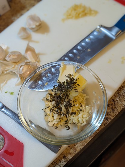 A cutting board with a large knife, some minced garlic, some garlic cloves, and a small glass bowl on it. In the glass bowl is butter, more minced garlic, and minced thyme.