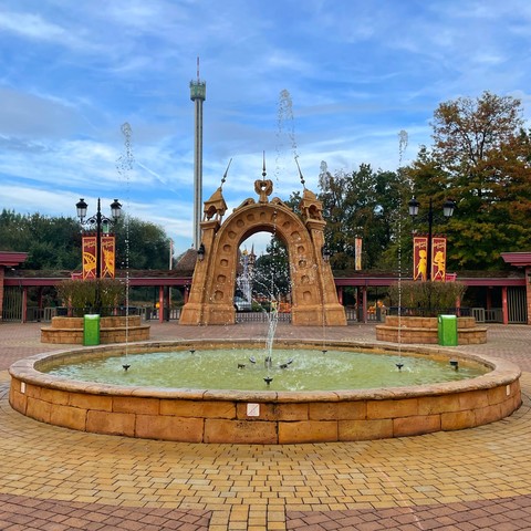 Photo of entrance to Holiday Park, Germany. Fountains in the foreground with an entrance arch behind. In the background, the Anubis Drop Tower is visible.