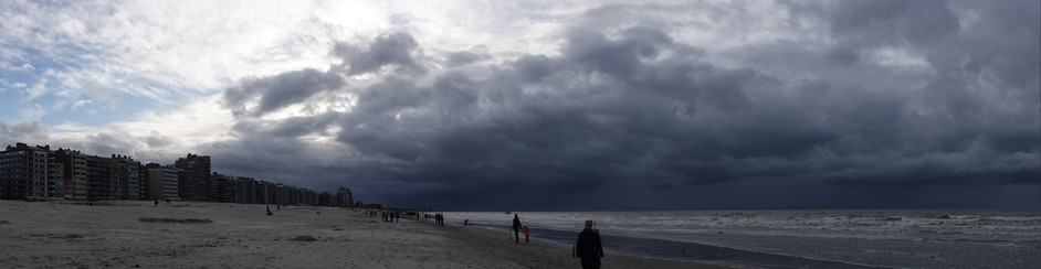 a wide panorama picture with some blue sky on the left but mostly very dark clouds, above the sea and beach