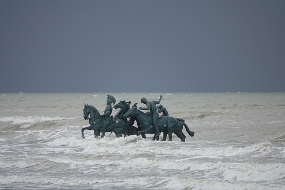 a sculpture consisting of men on horses, the sculpture was placed in te sea (to be consumed by the sea). The picture is taken with the sculpture surrounded by rough waves, beneath a dark grey sky