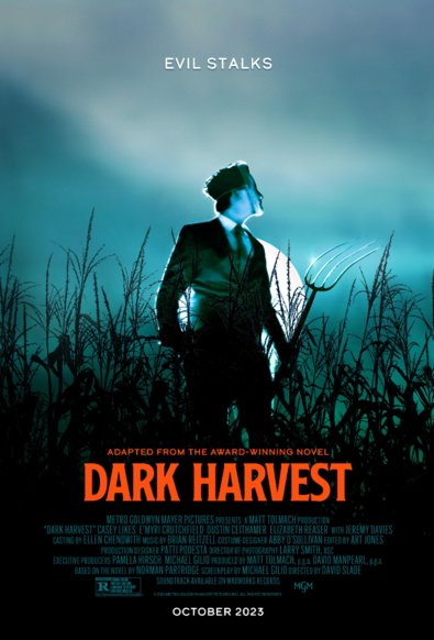 Film poster of the film "Dark Harvest". A young man in a dark suit with a white shirt and tie stands in front of a blue sky at night. He is wearing a distorted mask, holding a pitchfork and standing in a cornfield. He looks stressed and ready to attack or defend himself. Underneath in red lettering is the film title. 