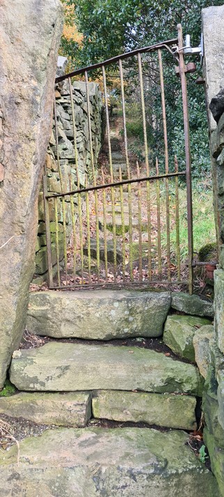 An old rusty metal gate, on top of three or four big stone steps. Beyond the gate, the steps continue up the hill into the distance alongside a traditional drystone wall. 