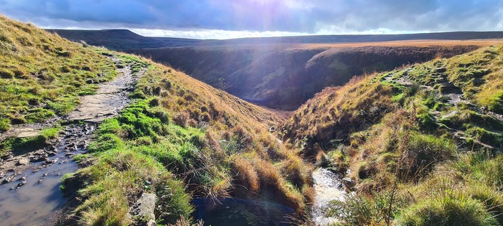 A moorland scene in autumn. Overlooking a "clough" - a valley formed by a stream, which feeds into a large valley/stream in the near distance. The foreground is lit by bright sunshine but the hills in the distance are dark, under the shadow of a heavy grey cloud.