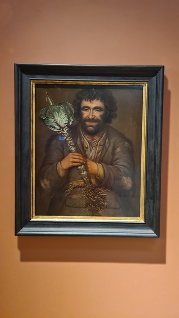 A man smiling out from an oil painting, he holds a big green white stick of kale over his right shoulder, smiling eyes and face, dark brown bushy hair, eyebrows and beard . He wears a threadbare jacket with elbows worn through, old shirt and belt underneath 