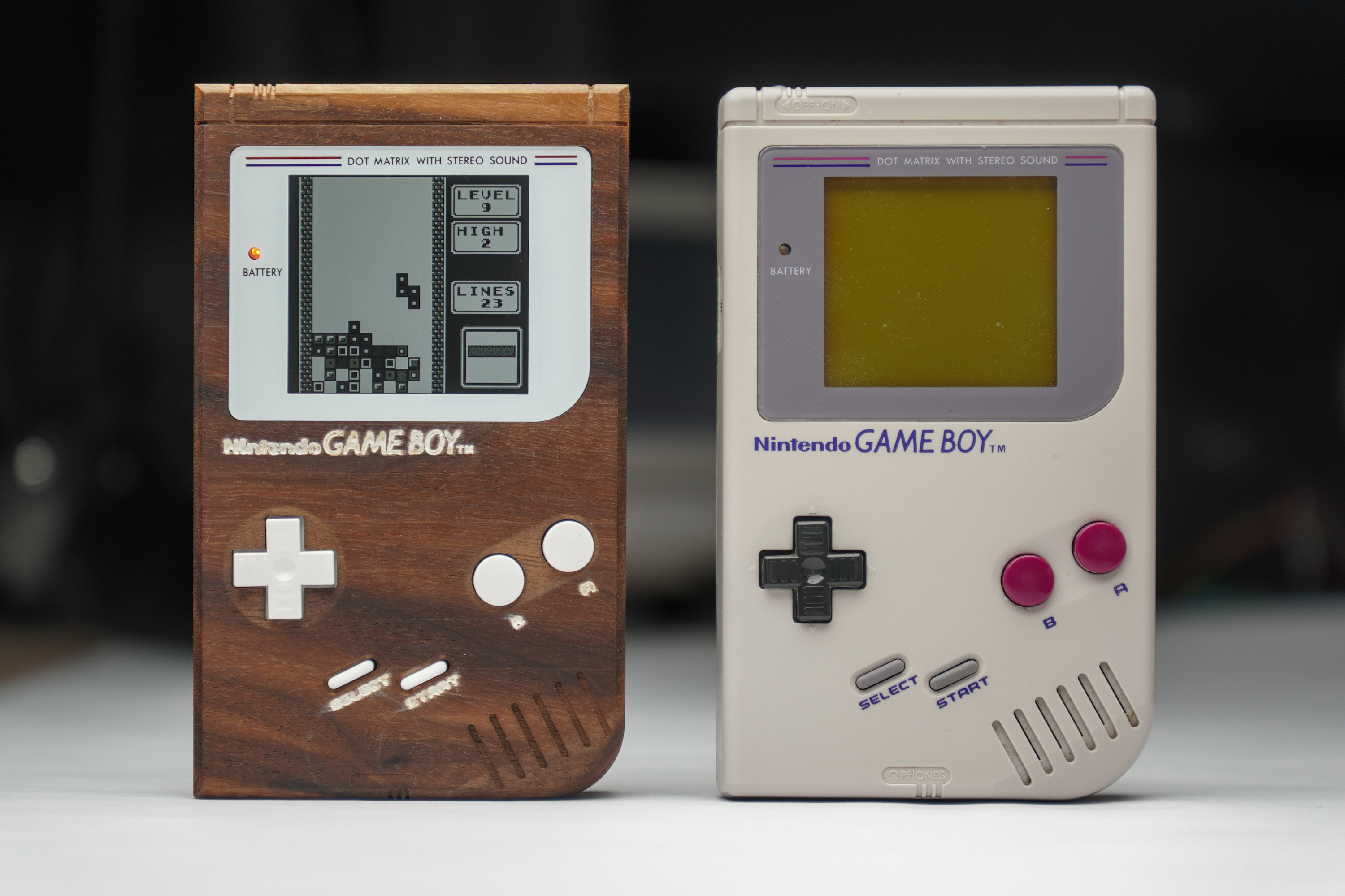 Photo of the dark brown wooden Game Boy with white buttons and white screen bezel standing next to an original Game Boy with a light gray injection mold case, a gray screen bezel, black D-pad and red A and B buttons.