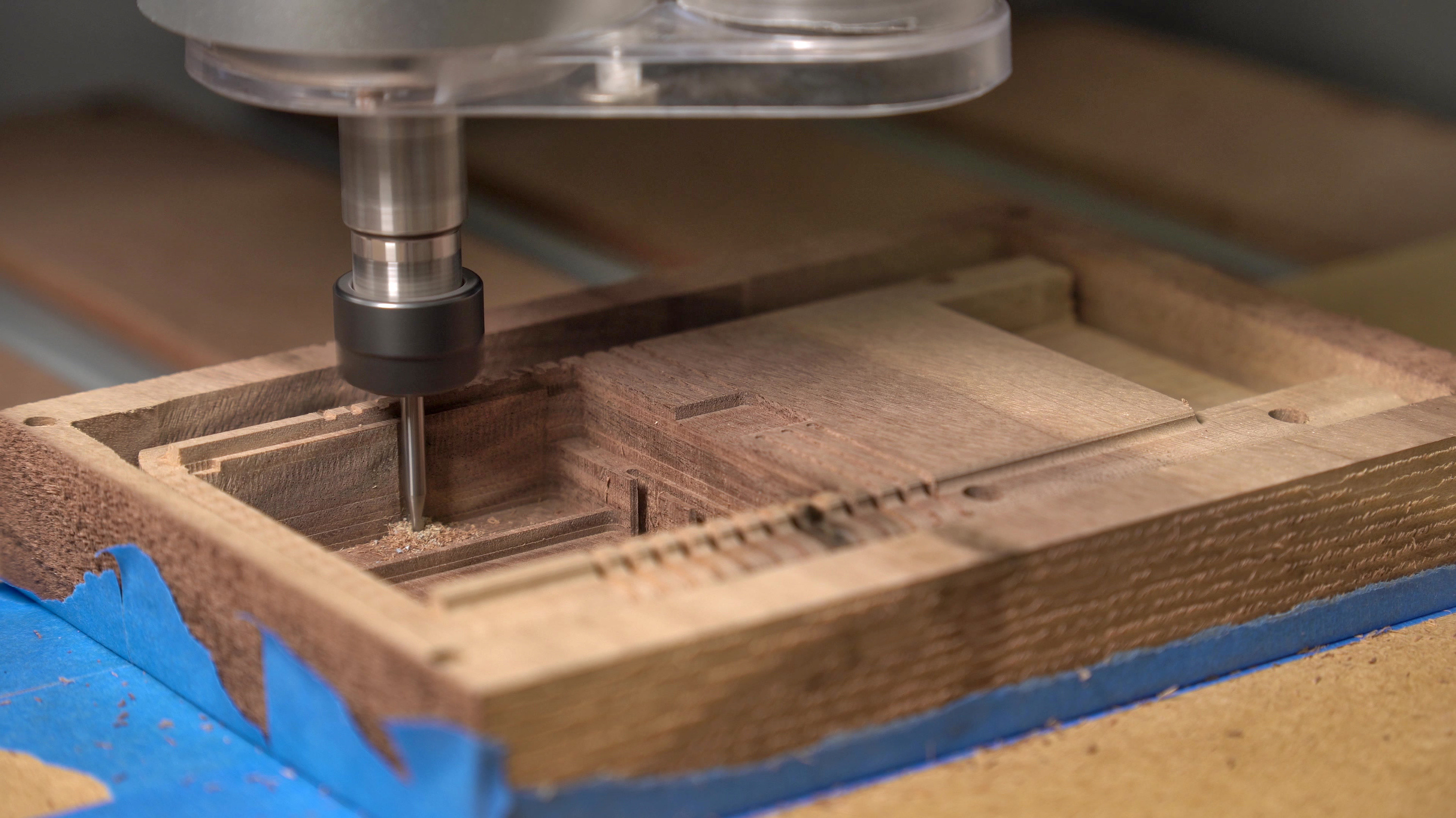 Photo of the CNC machining process. A block of wood on the wooden bed of a CNC router is already showing the recognizable design of the backside of a Game Boy. A spinning fine end mill is drilling into a part of the battery compartment. The wood is noticably brighter than the finished Game Boy as it has not yet been oiled.