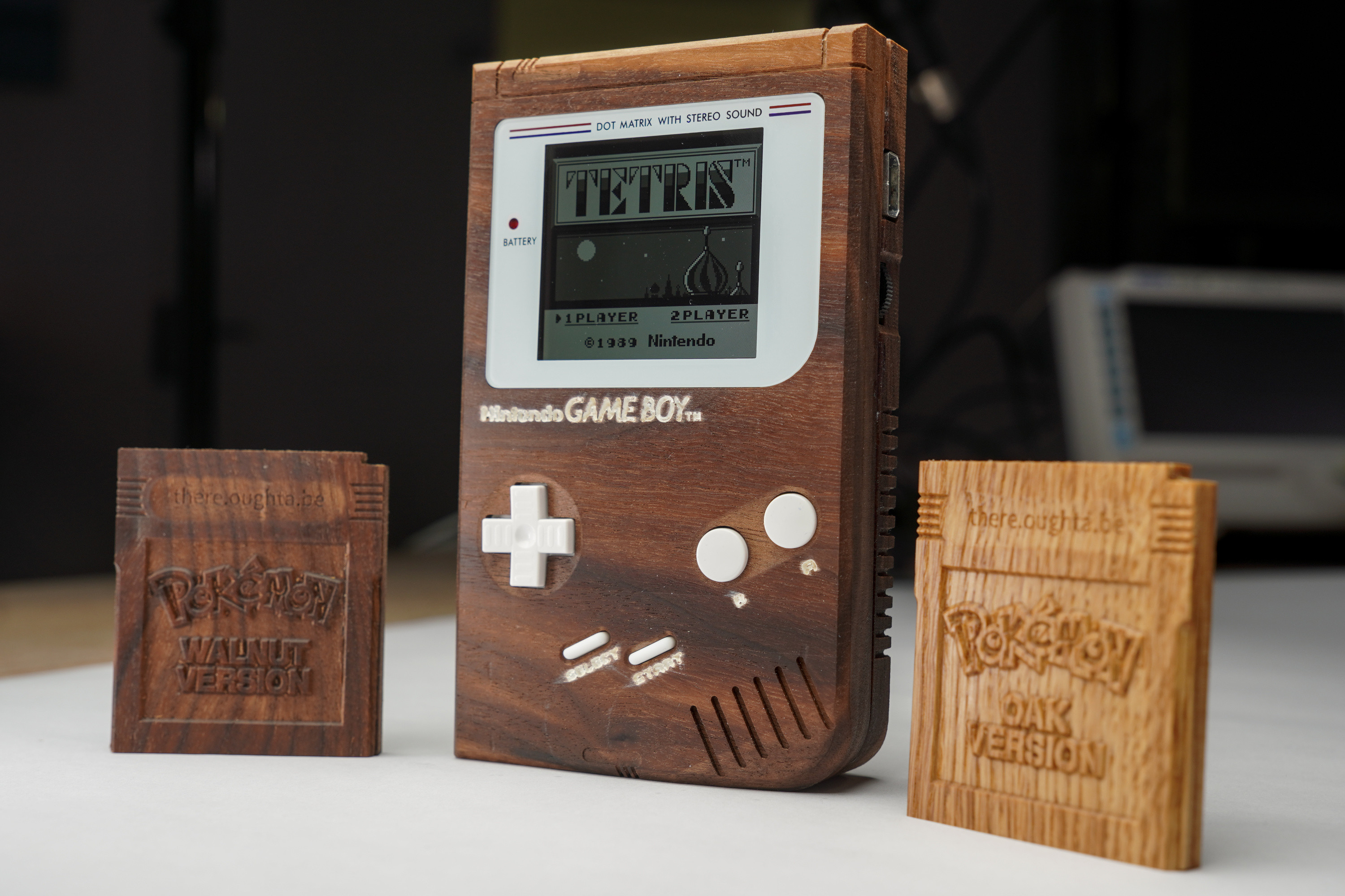 Photo of the dark brown wooden Game Boy with white buttons and white screen bezel ist standing on a white surface with a dark blurred backdrop. It shows the title screen of Tetris. To the left and right are wooden Game Boy cartridges. The left one has the same color as the Game Boy and has a carved label reading "Pokemon Walnut version". The other one is made of brighter wood and reads "Pokemon Oak version".