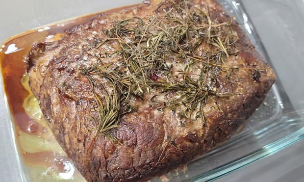 Farbfoto: Ein großes Stück Fleisch liegt in einer Glasschale. Auf dem Fleisch liegen Kräuter, in der Schale hat sich Fleischsaft gesammelt. Das Fleisch ist außen kräftig angebraten. 