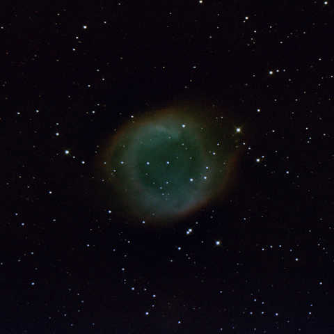 The Helix Nebula, seen here as a hazy, roundish mass of gasses sloughed off by the white dwarf star at its center. The middle is greenish blue, becoming yellowish toward the outside, fringed in red. Background is black with a moderate amount of small stars.