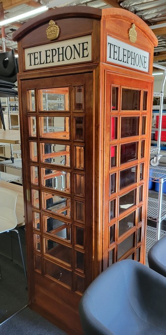 Stained wooden version of the red telephone box at an office furniture liquidation store. At the top of each side is the Crown, a symbol of the British government. The word Telephone appears below the crown on each side in large black block letters on an off-white background.