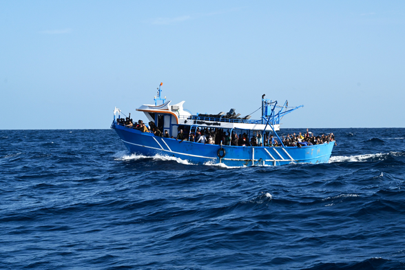 Ein überfülltes blaues Fischerboot in Fahrt auf offener See. Es ist Tag, der Himmel klar. Die See leicht unruhig.