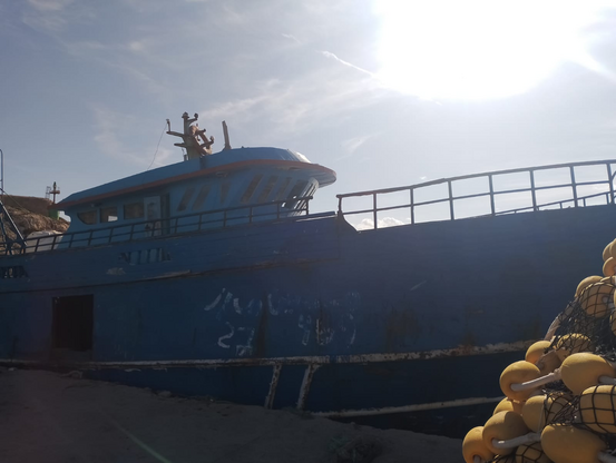 Ein leeres blaues Fischerboot im Gegenlicht am Kai. Am Rumpf eine verblassende Markierung mit Kreide. Eine 27 ist noch zu erkennen.