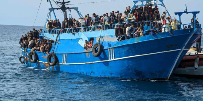 Ein überfülltes blaues Fischerboot bei ruhiger See auf dem Meer. Im Hintergrund schaut der Bug einer Guardia Costiera CP hervor.