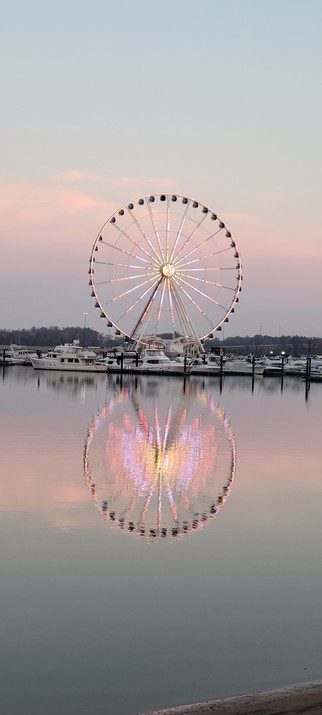 Lit ferris wheel reflected in the water with boats at its base and pink clouds behind