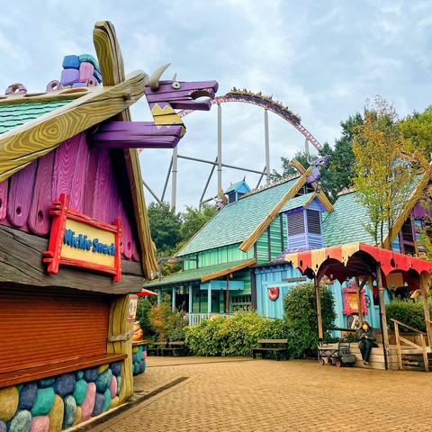 Photo of Expedition GeForce cresting an airtime hill. Photo taken at an angle such that the track goes through the mouth of a cartoonish purple beast at the top of a closed  “Wickie Snack” kiosk. 