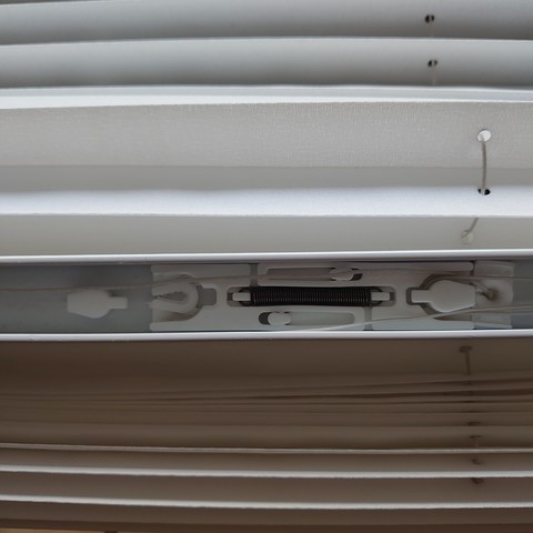 Foto av skyvemekanismen inni en plissé-gardin. På hver side av mekanismen er det en form for trinse, bare uten hjul. Det er istedet et dråpeformet plaststikke som tauet skal gli over. Den venstre trinsen er ødelagt og i to deler på grunn av at tauet har gnagd seg igjennom plasten.
