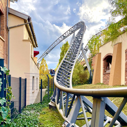 Photo of track of Karacho coaster diving down towards the camera between two buildings. A train is just visible on the brake run in the background. The sky is a bright mixture of blue sky and clouds.
